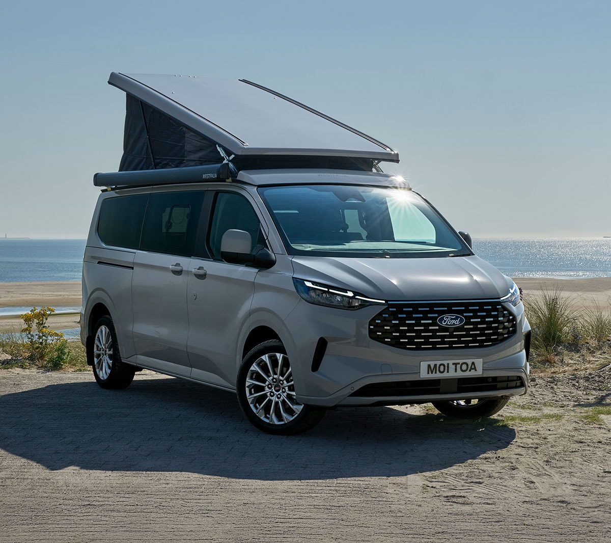 Stříbrný Ford Transit Custom Nugget s otevřenou střechou zaparkovaný na pláži u moře.
