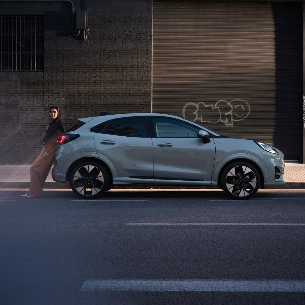 Žena se opírá o zaparkovaný šedý Ford Puma v městském prostředí.