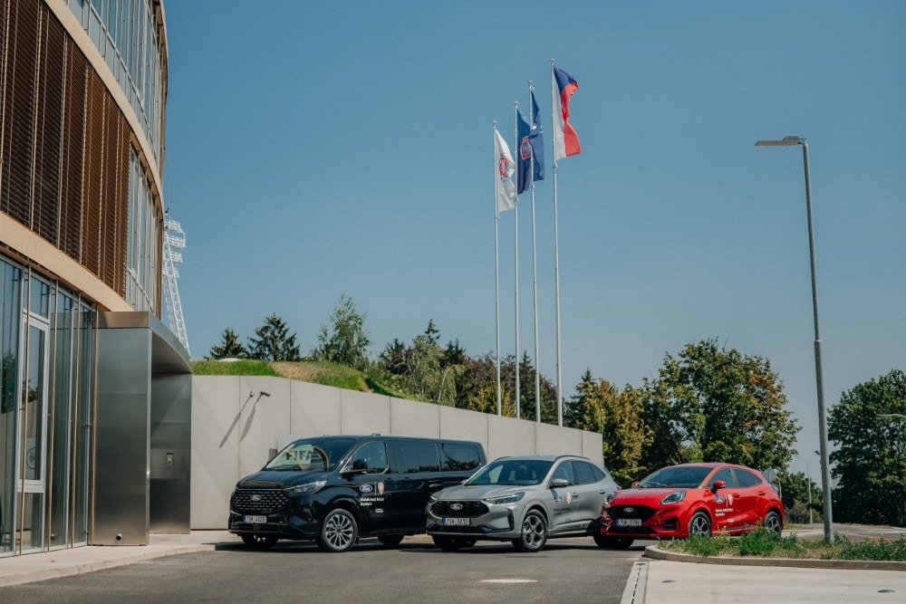 Černý Ford Tourneo Custom, šedý Ford Kuga a červený Ford Puma jsou zaparkovány před moderní budovou s vlajkami.
