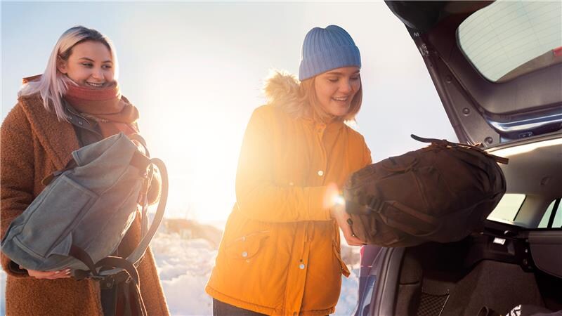 Dva lidé nakládají batohy do otevřeného kufru auta za slunečného zimního dne.