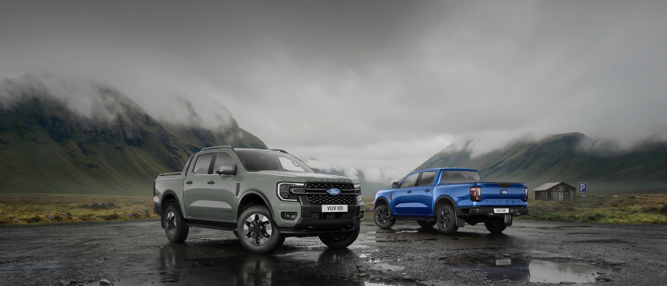 Dva Fordy Ranger, šedý a modrý, zaparkované na mokré ceste s horami v pozadí.