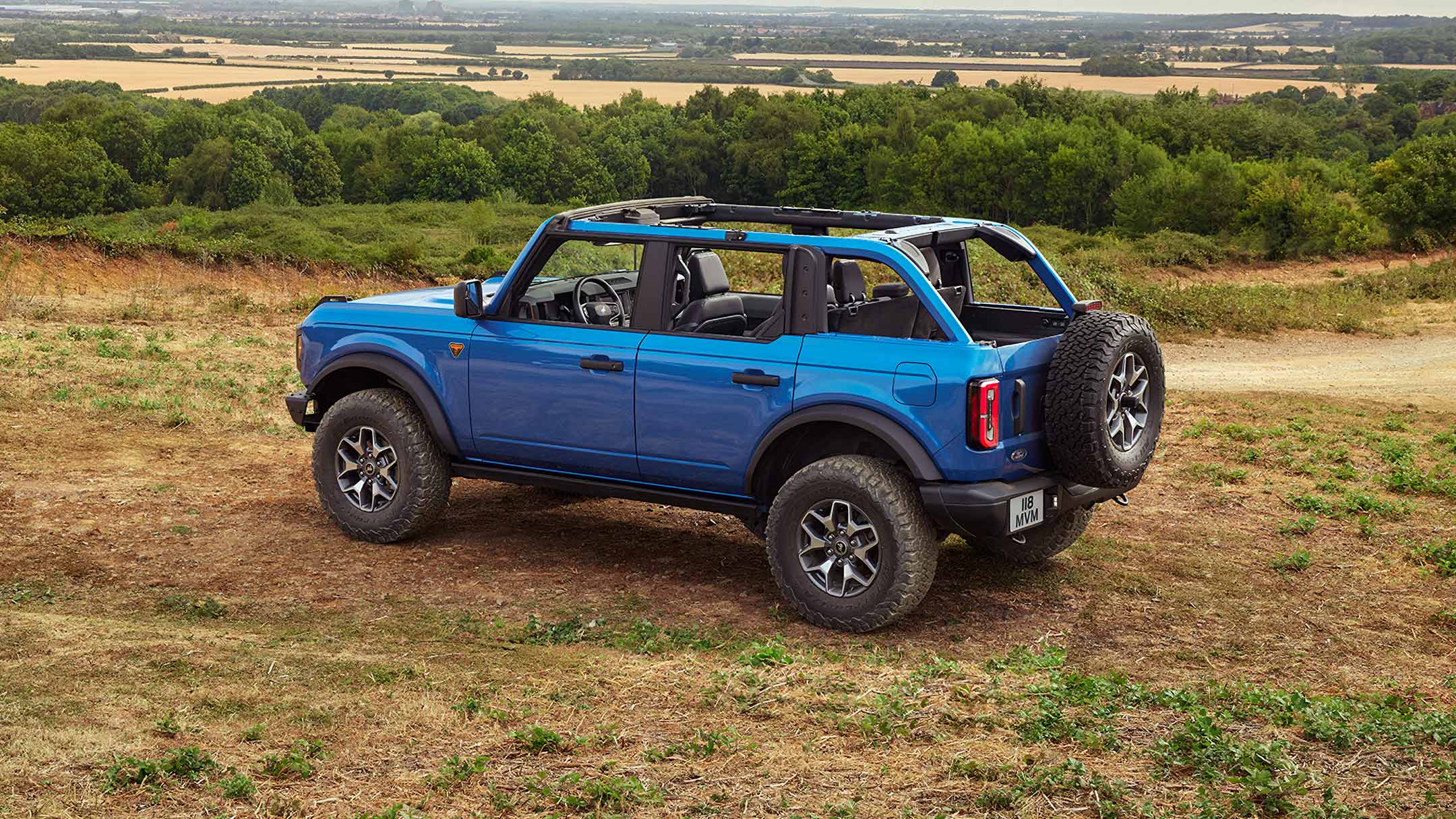 Modrý Ford Bronco s odnímatelnou střechou a dveřmi, zaparkovaný na prašné terénní cestě.