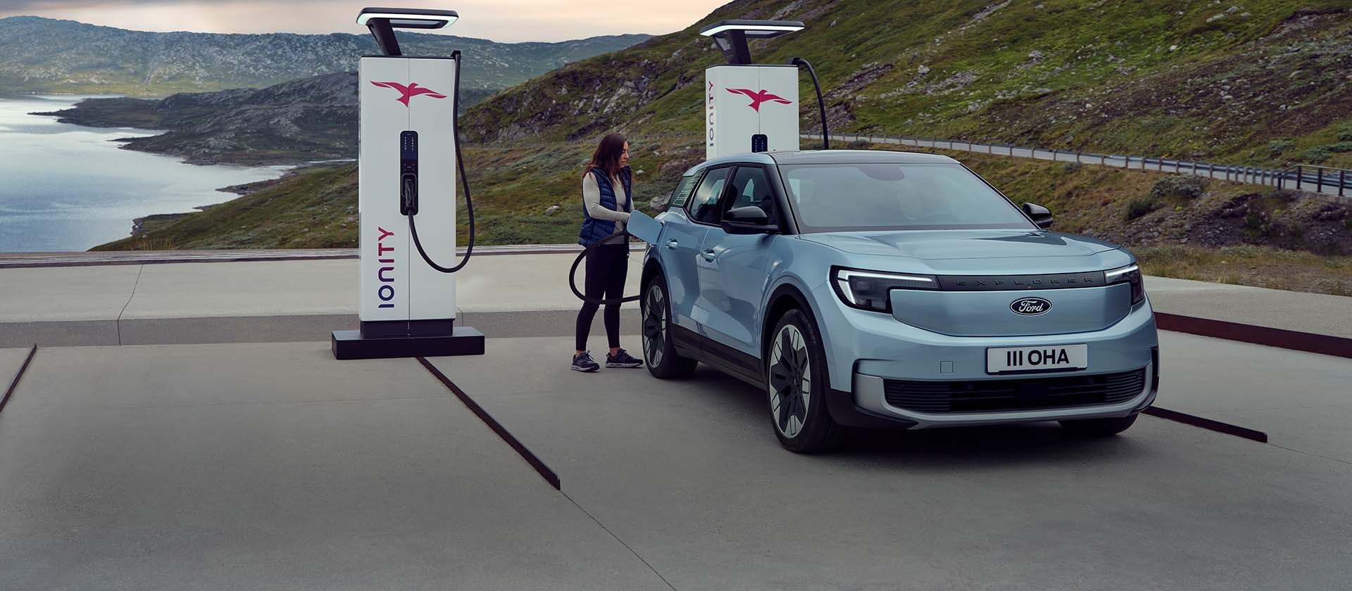 Žena zapojuje modrý, plně elektrický Ford Explorer pomocí kabelu do veřejné nabíjecí stanice. Auto je zaparkované u hor a vody.