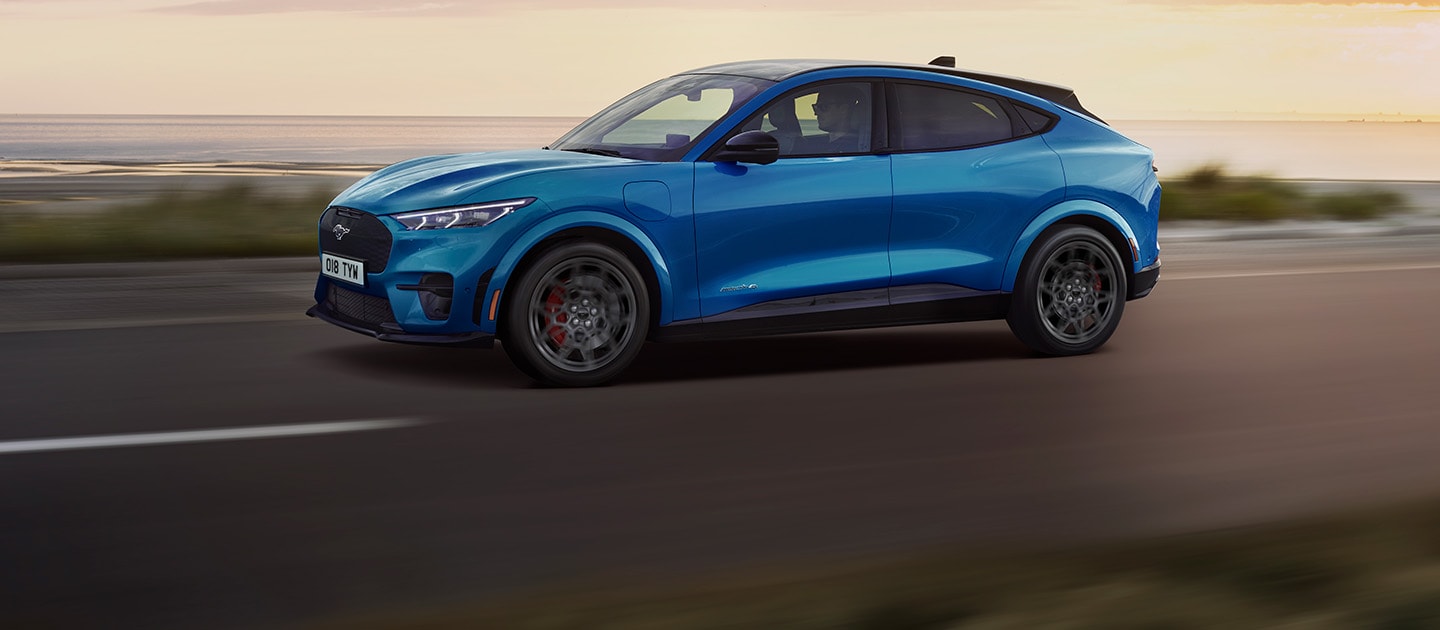 Modrý elektrický Ford Mustang Mach-E jedoucí po pobřežní silnici při západu slunce.