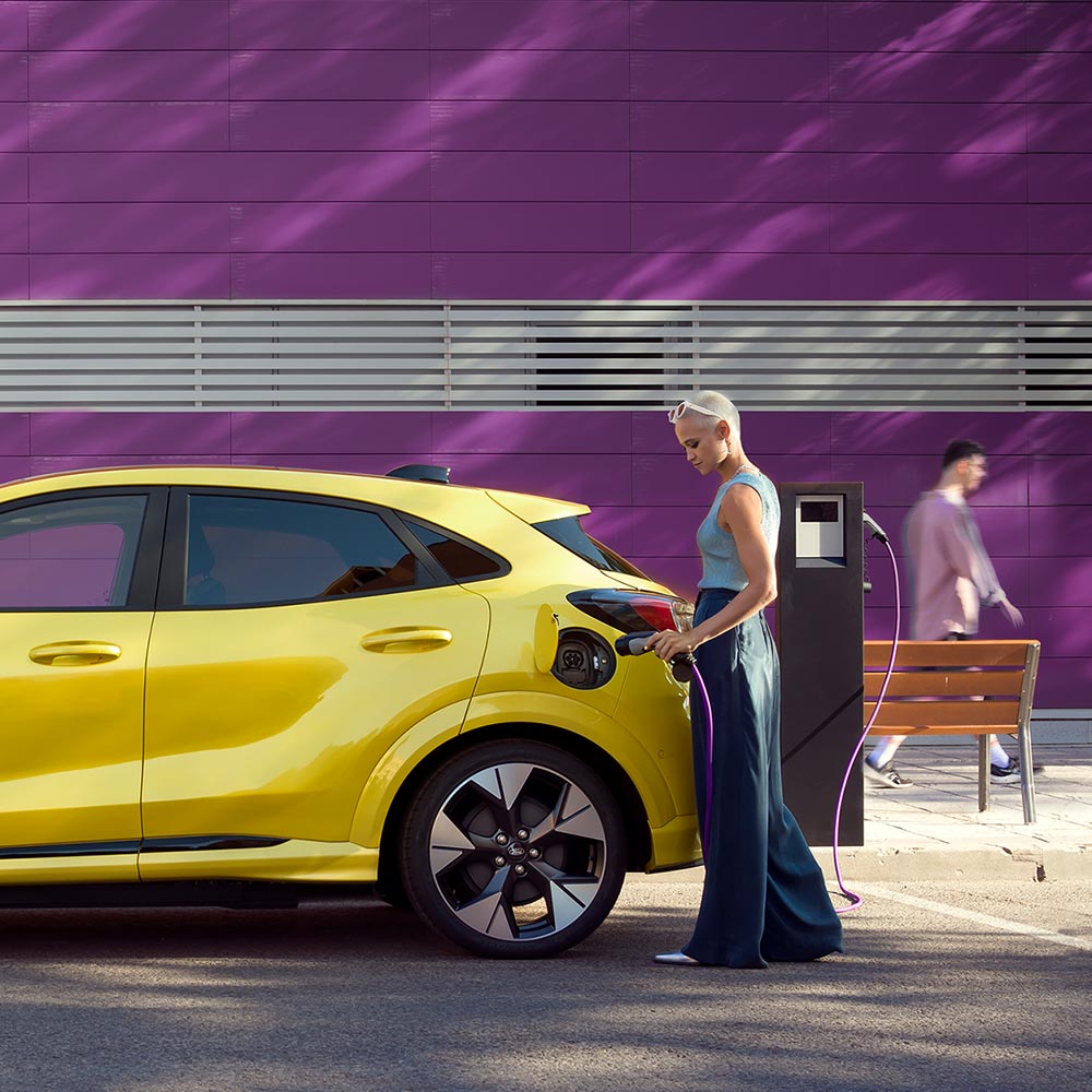 A driver plugging a yellow Ford Puma Gen-E into a public charging station for easy EV charging.