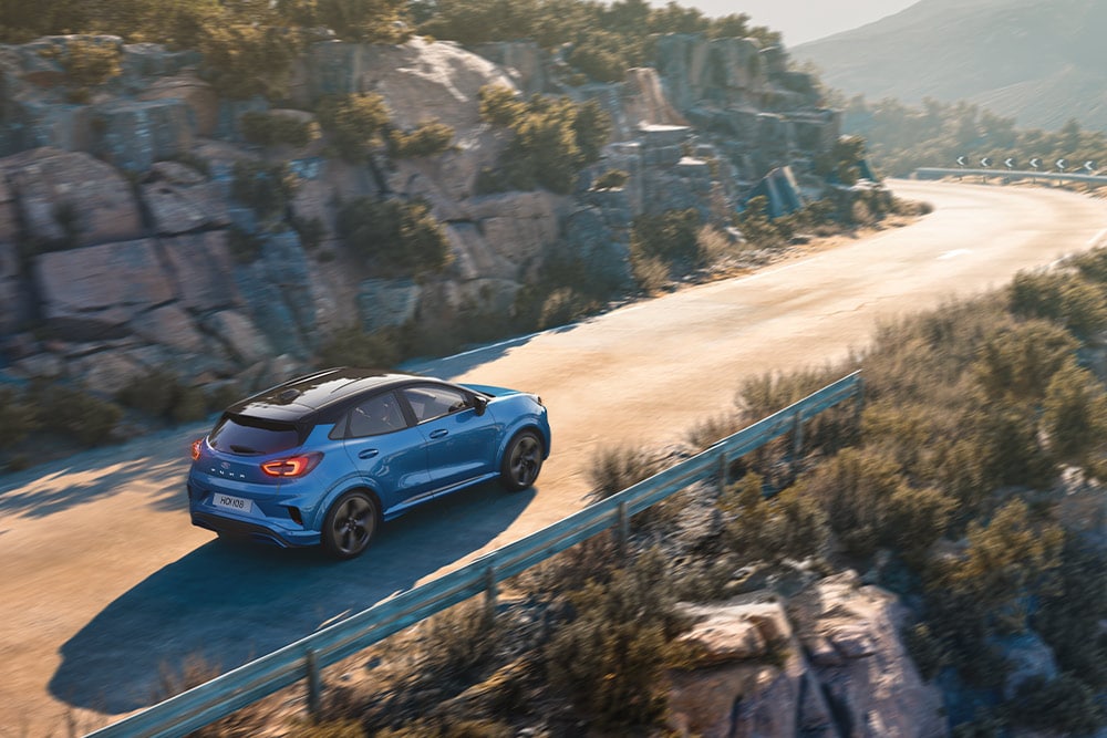 Modrý Ford Puma Gen-E jede po klikaté horské silnici.