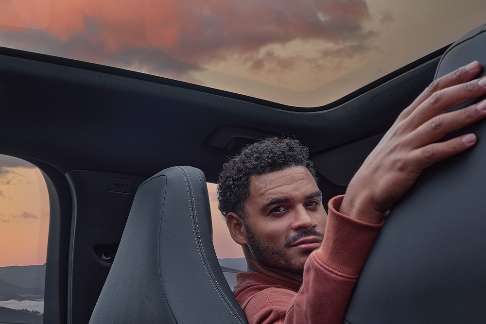 A man sitting in a car at sunset with his hand resting on the back of one of the seats.