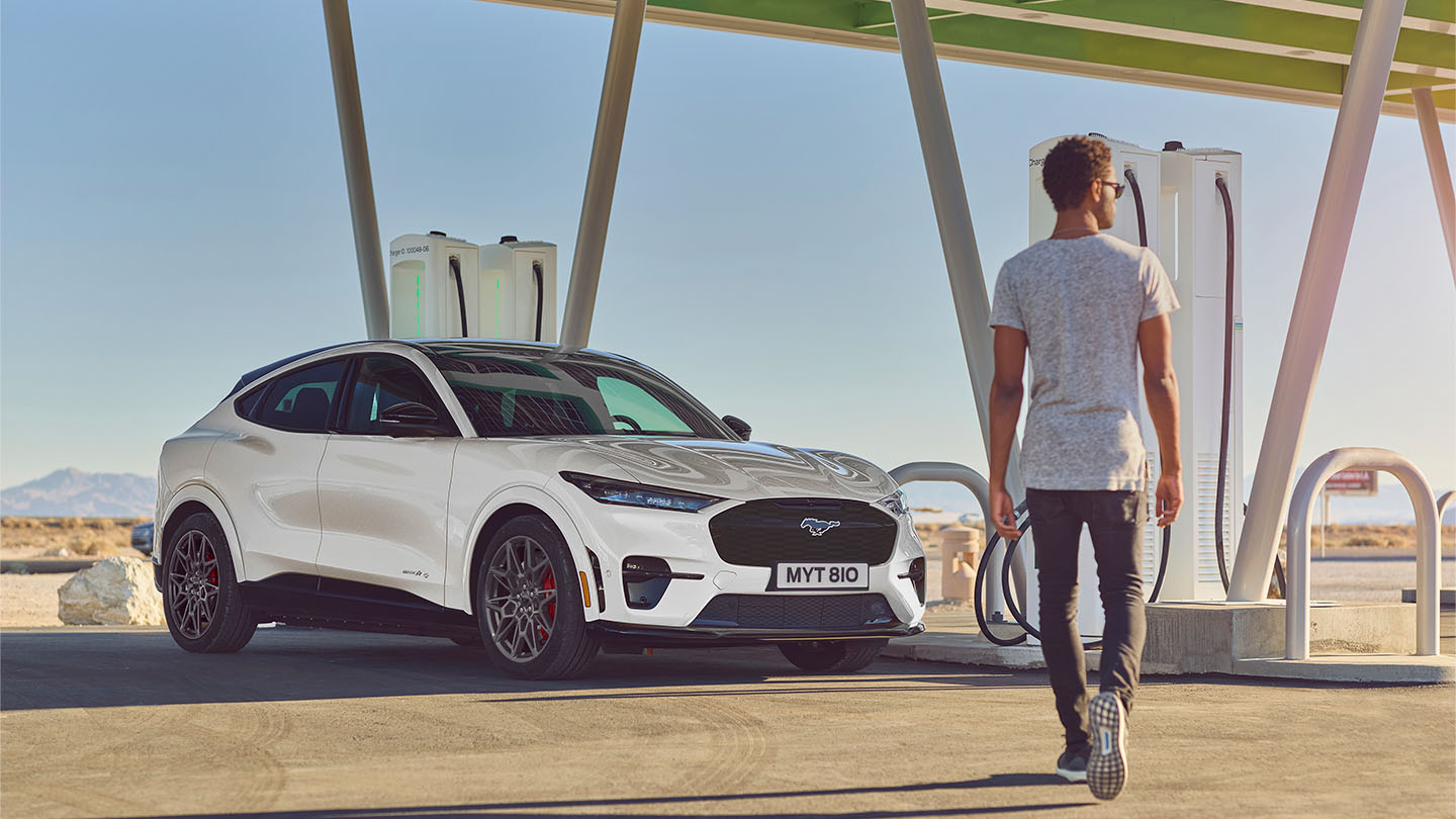 Bílý Ford Mustang Mach-E zaparkovaný u venkovní veřejné nabíjecí stanice. Muž v bílém tričku kráčí k vozu.