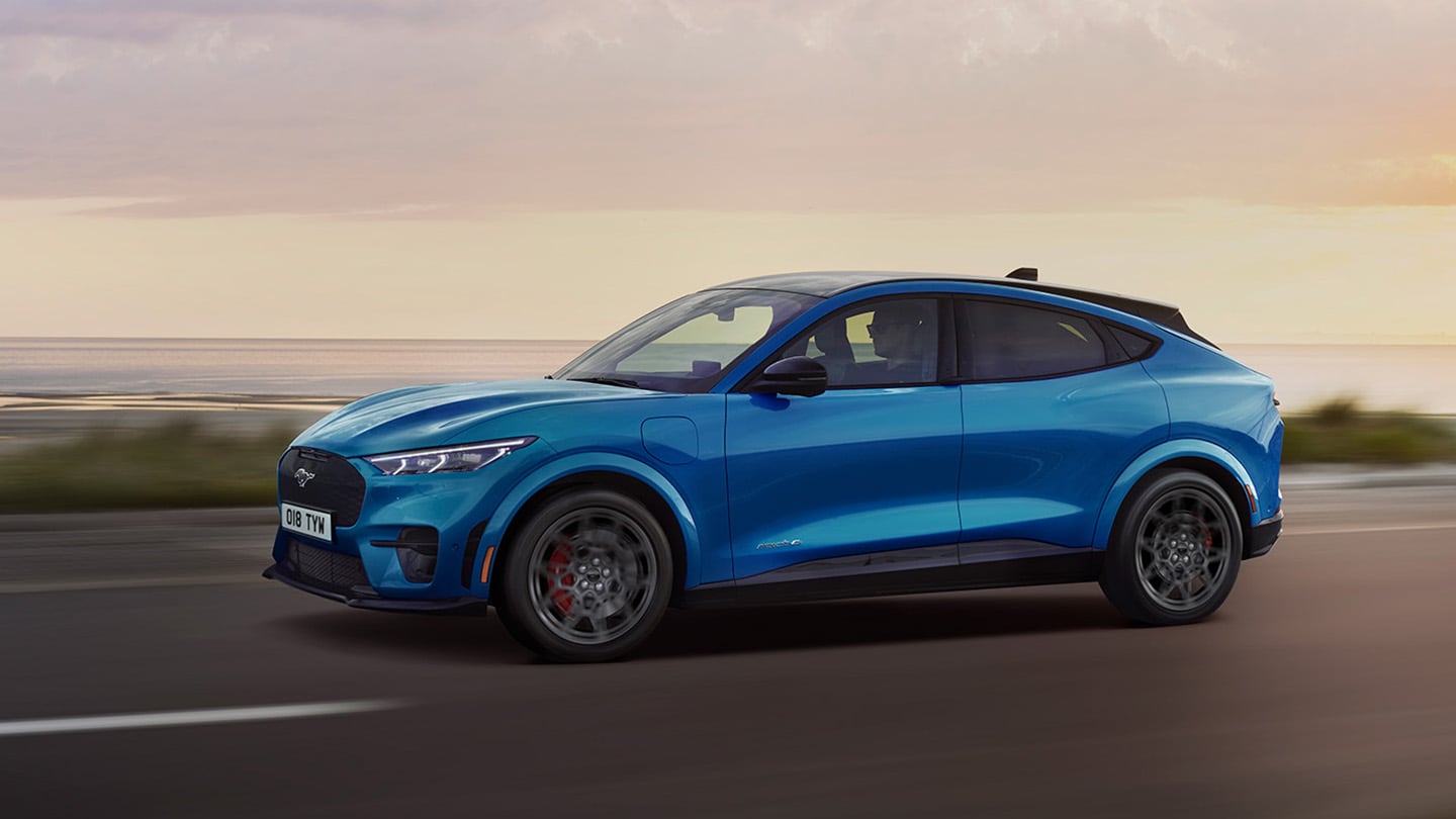 Boční pohled na modrý elektrický Ford Mustang Mach-E při jízdě za soumraku po silnici podél pobřeží.