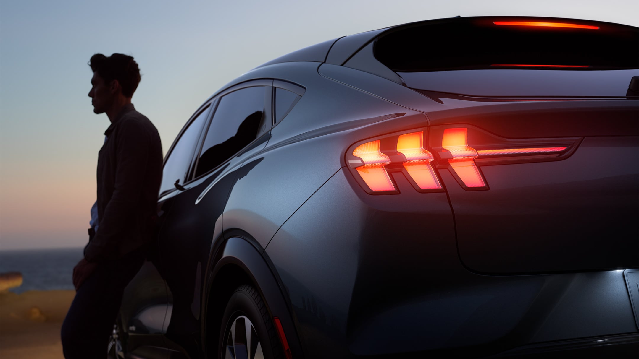 Muž opírající se o zbrusu nový Ford Mustang Mach-E během západu slunce