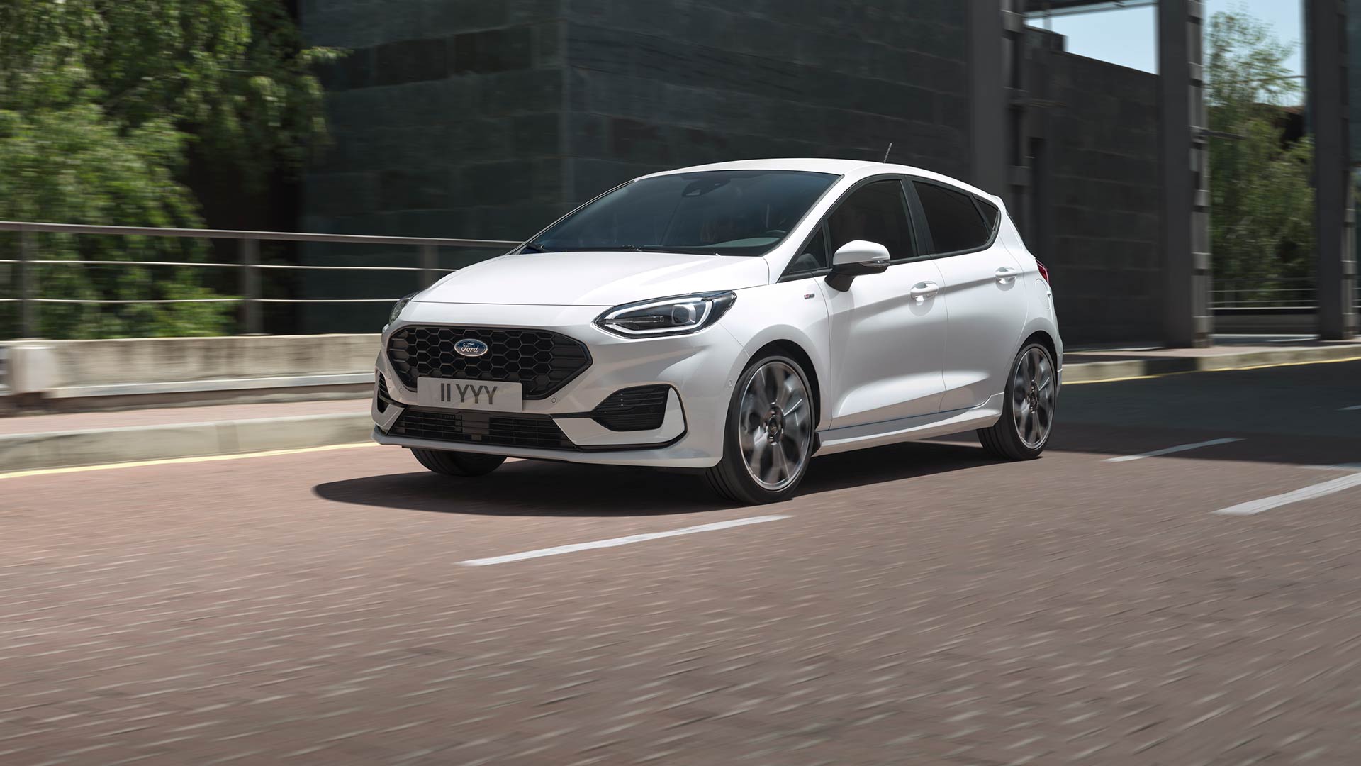 Bílý Ford Fiesta jedoucí po městské silnici lemované zelení a budovami.