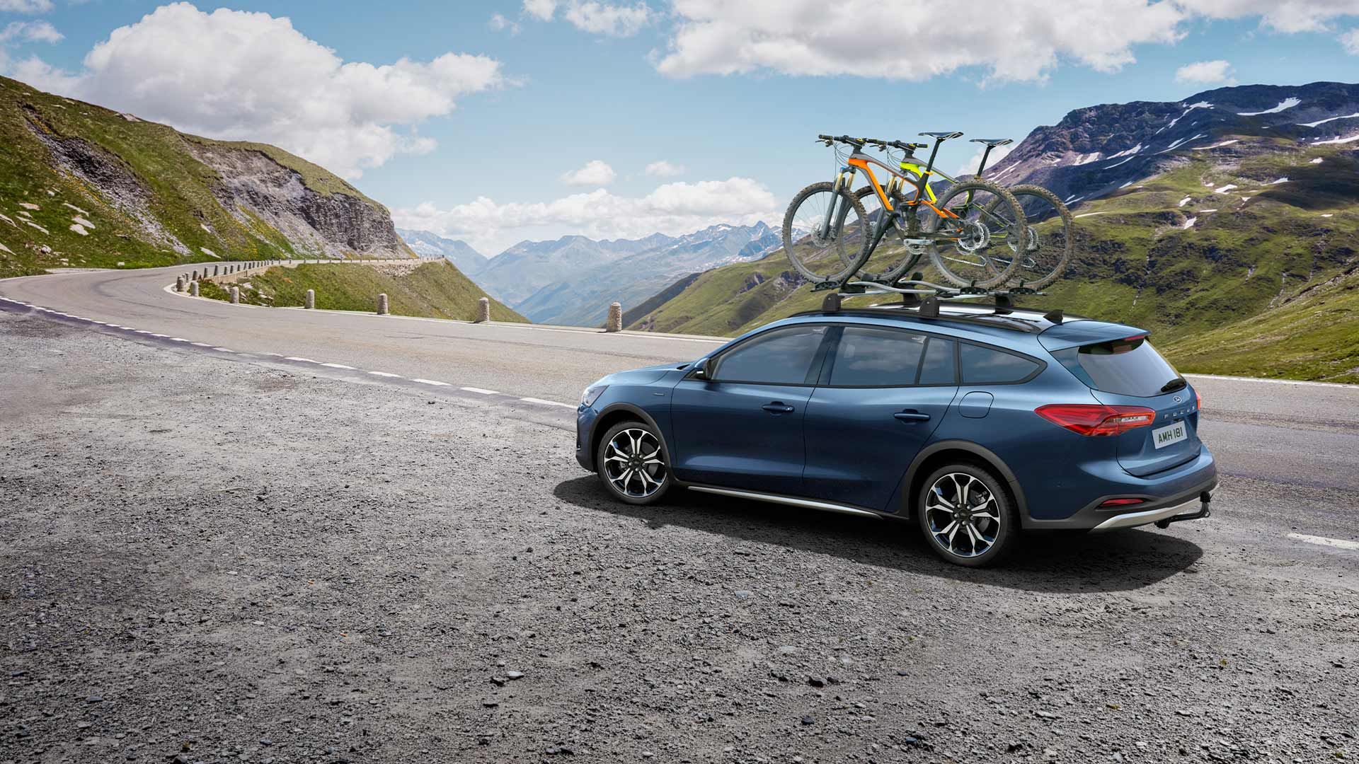 Modrý Ford Focus s jízdními koly na střeše jedoucí po malebné horské cestě.