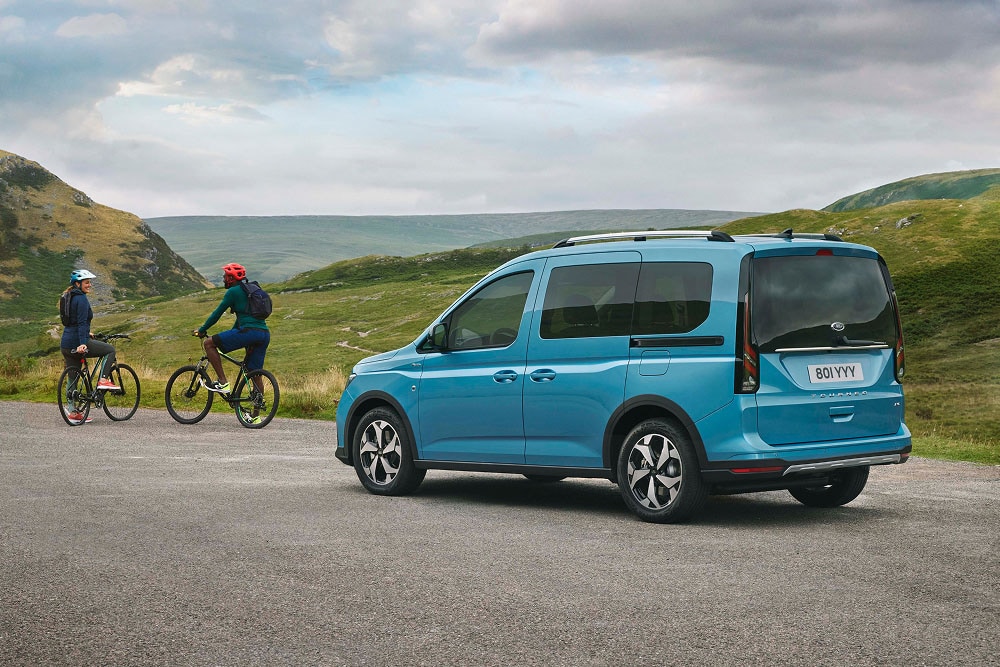Modrý Ford Tourneo Connect stojí na silnici v zelené kopcovité krajině a poblíž jsou dva lidé na jízdních kolech.
