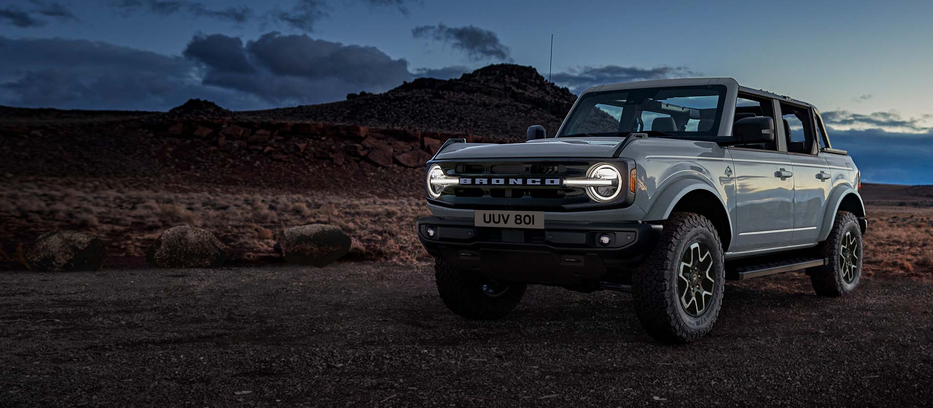 Boční pohled na Ford Bronco v šedé barvě, jak stojí zaparkovaný za soumraku ve skalnaté krajině.
