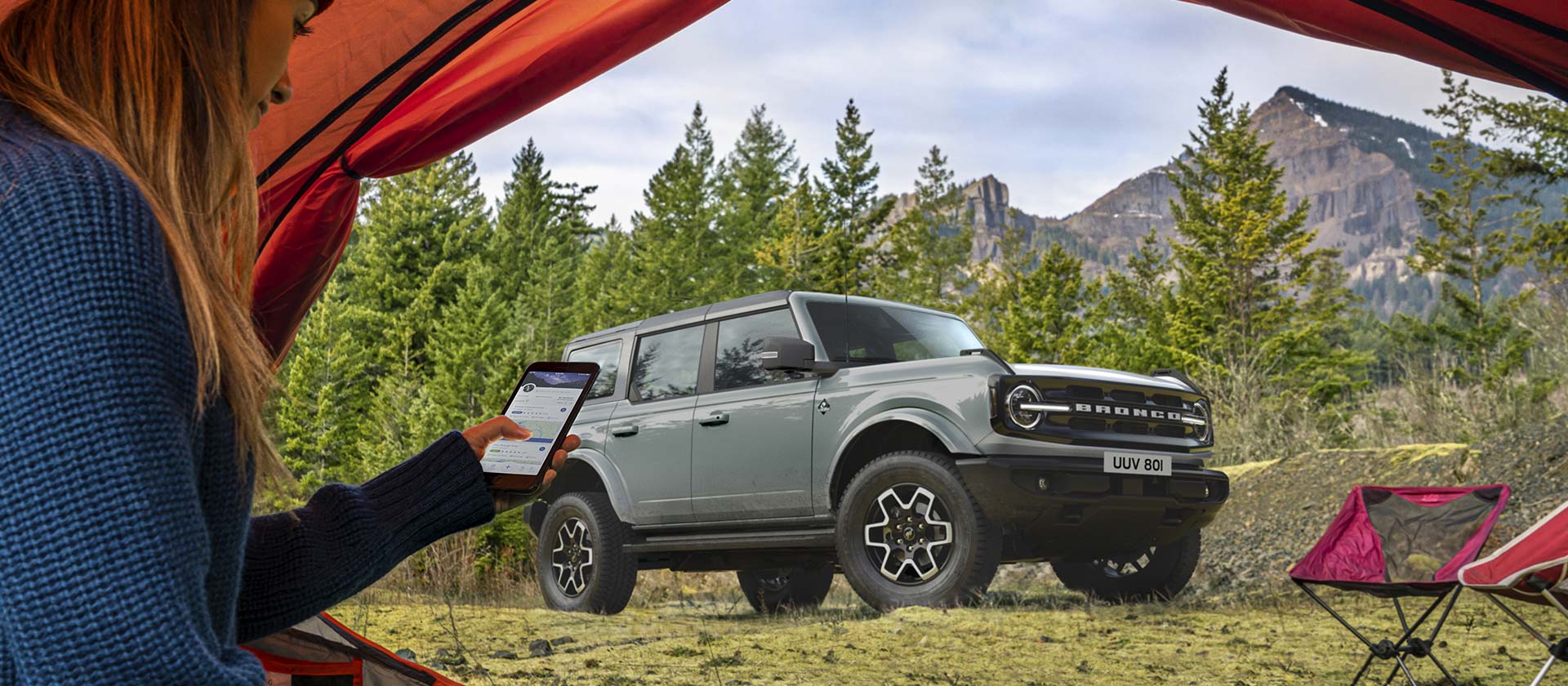 Pohled na Ford Bronco v horském kempu, před ním je osoba s telefonem ve stanu.