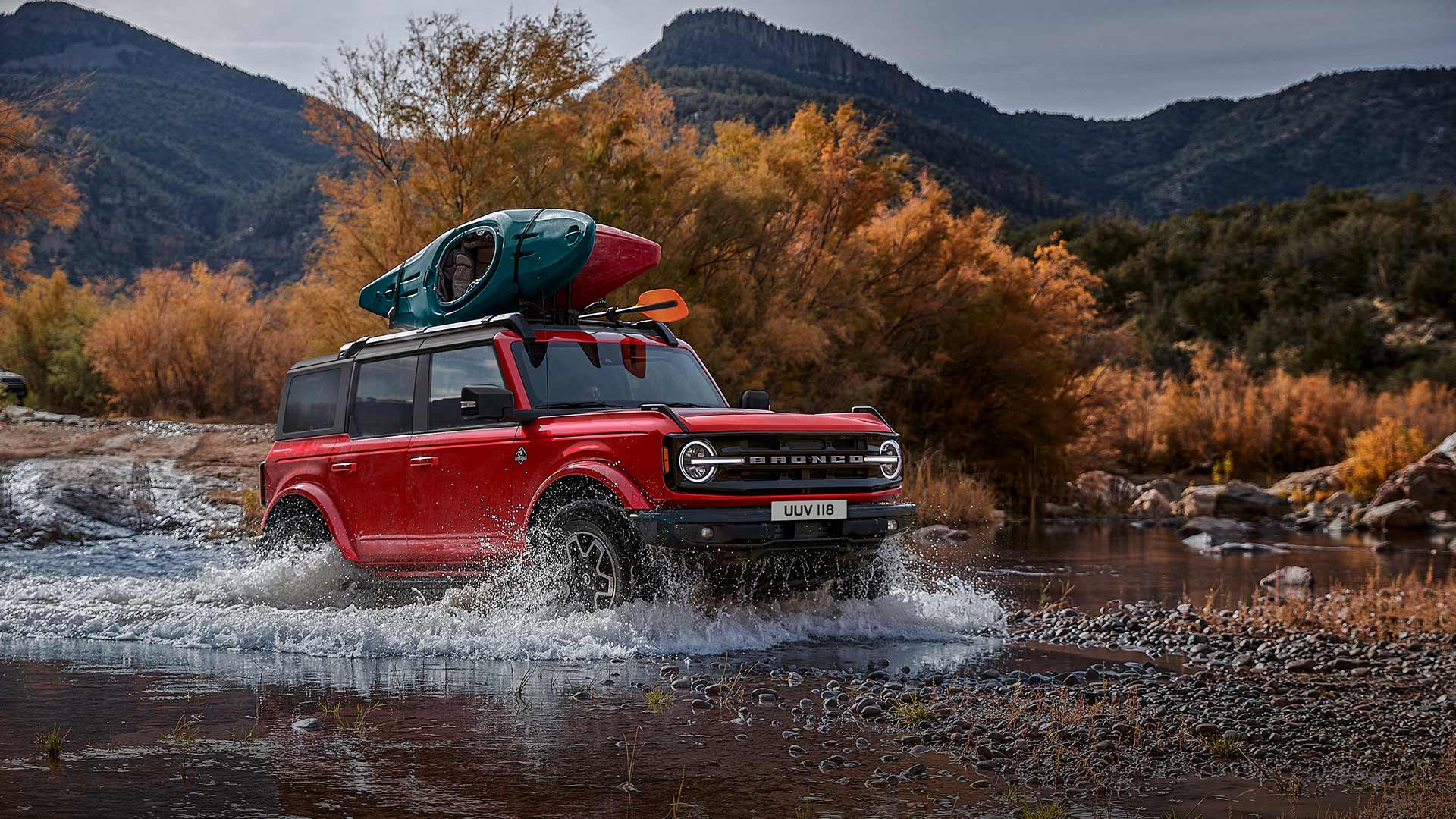 Boční pohled na červený Ford Bronco, který projíždí mělkým brodem v horské krajině.