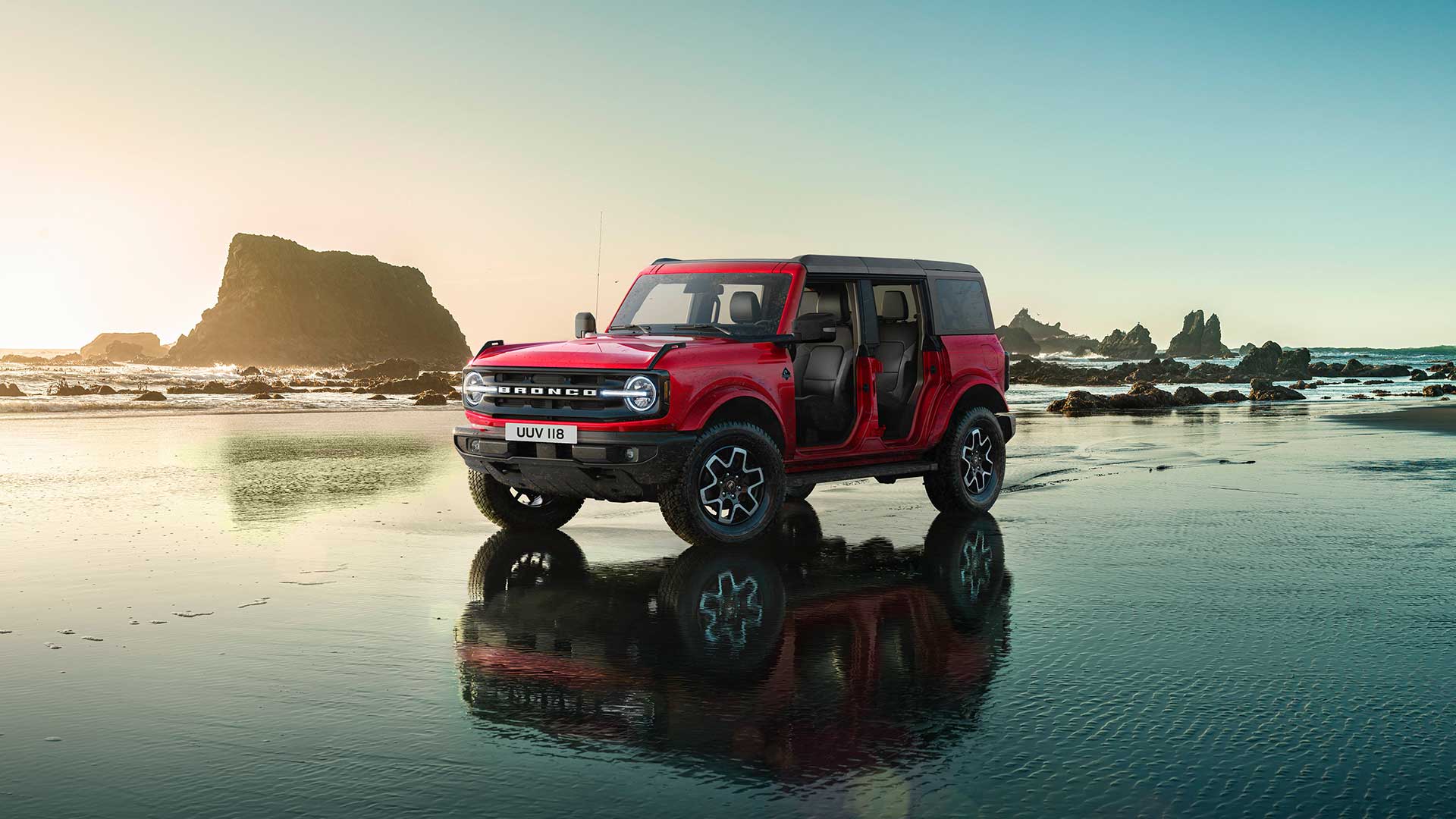 Boční pohled na červený Ford Bronco s odstraněnými dveřmi, jak stojí zaparkovaný na mokrém písčitém břehu.