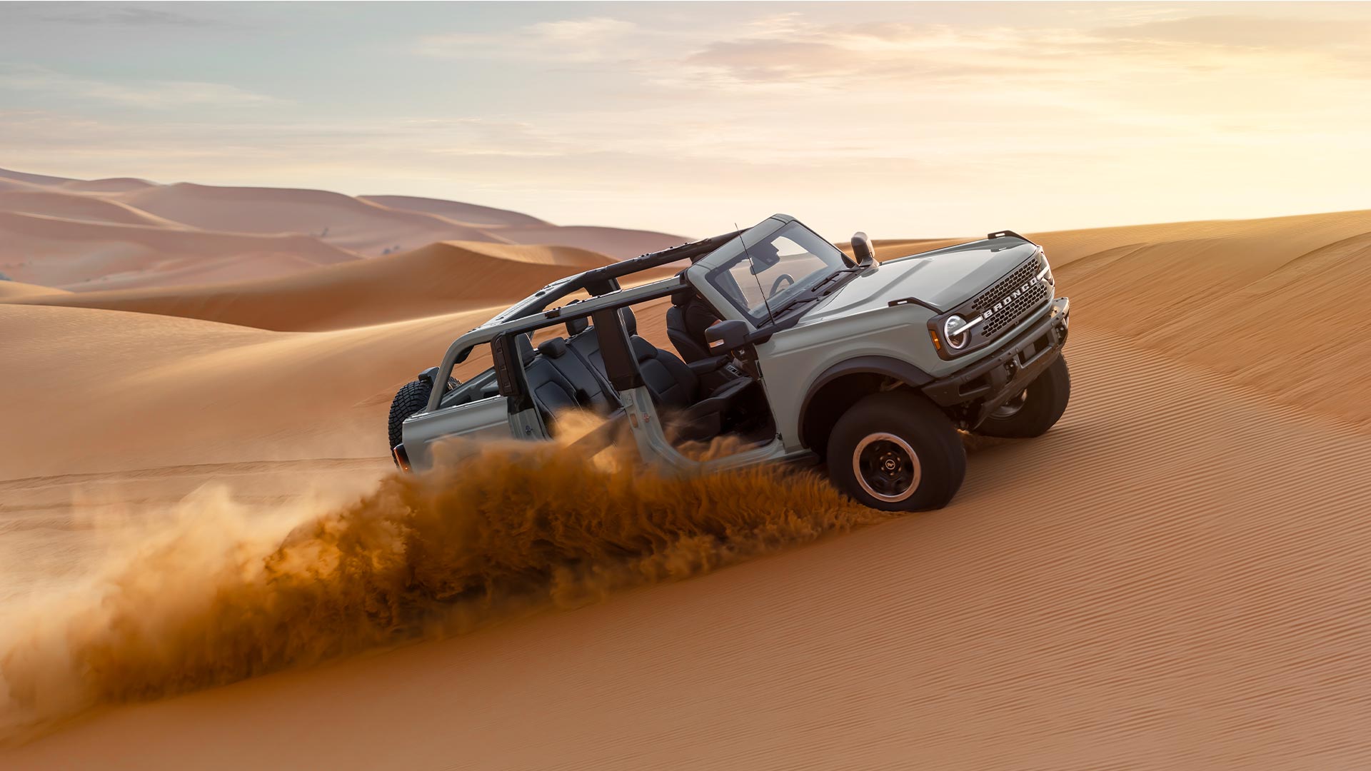 Boční pohled na šedý Ford Bronco s odstraněnými dveřmi a střechou, jak projíždí písečnou pouští.