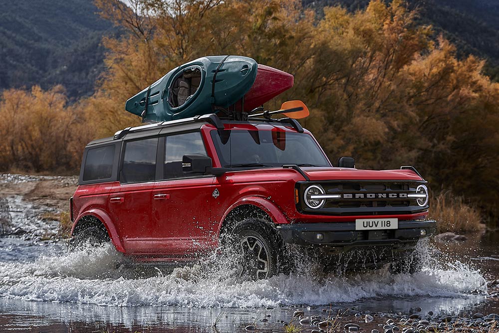 Boční pohled na červený Ford Bronco, který projíždí mělkým brodem v horské krajině.