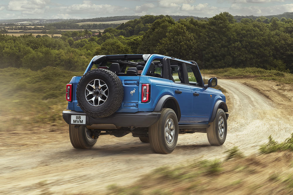 Pohled zezadu na modrý Ford Bronco projíždějící venkovskou krajinou.