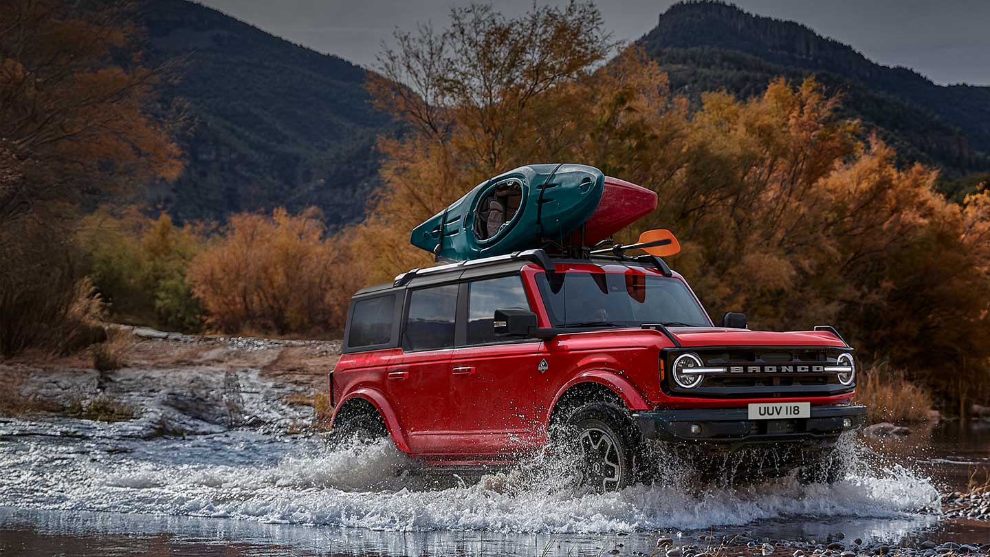 Boční pohled na červený Ford Bronco, který projíždí mělkým brodem v horské krajině.