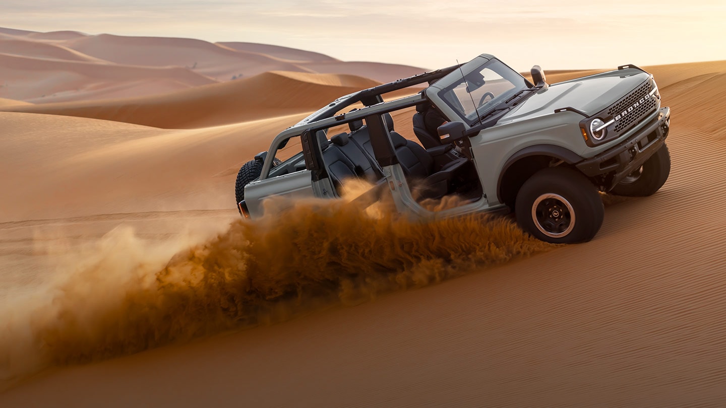 Boční pohled na šedý Ford Bronco s odstraněnými dveřmi a střechou, jak projíždí písečnou pouští.
