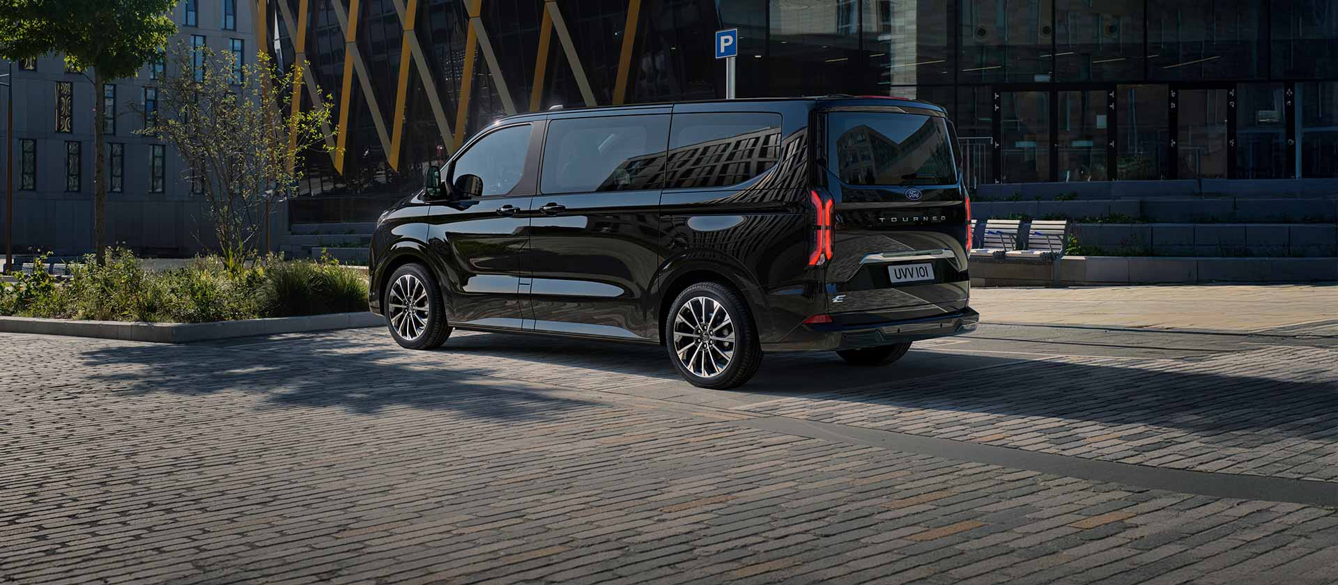 Před moderní budovou parkuje černý Ford E-Tourneo Custom při pohledu z boční a zadní strany.