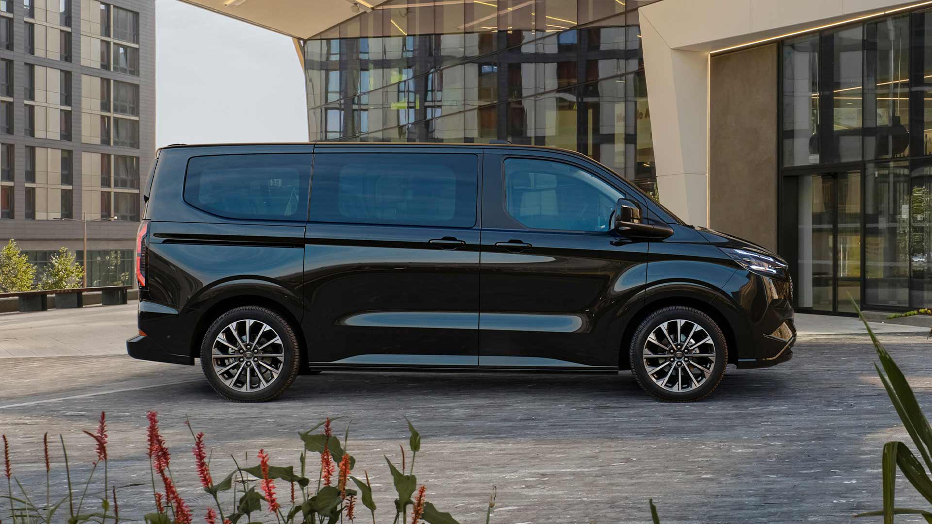 Před moderní budovou parkuje černý Ford E-Tourneo Custom.