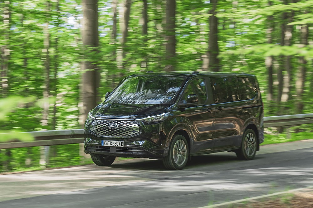 Černý Ford E-Tourneo Custom jedoucí po silnici lemované stromy.