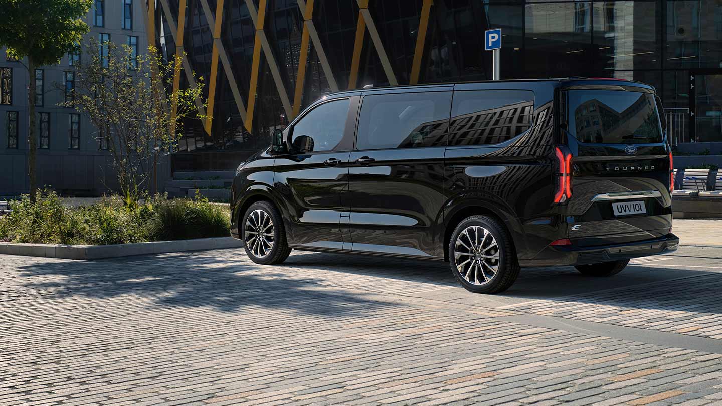 Před moderní budovou parkuje černý Ford E-Tourneo Custom při pohledu z boční a zadní strany.