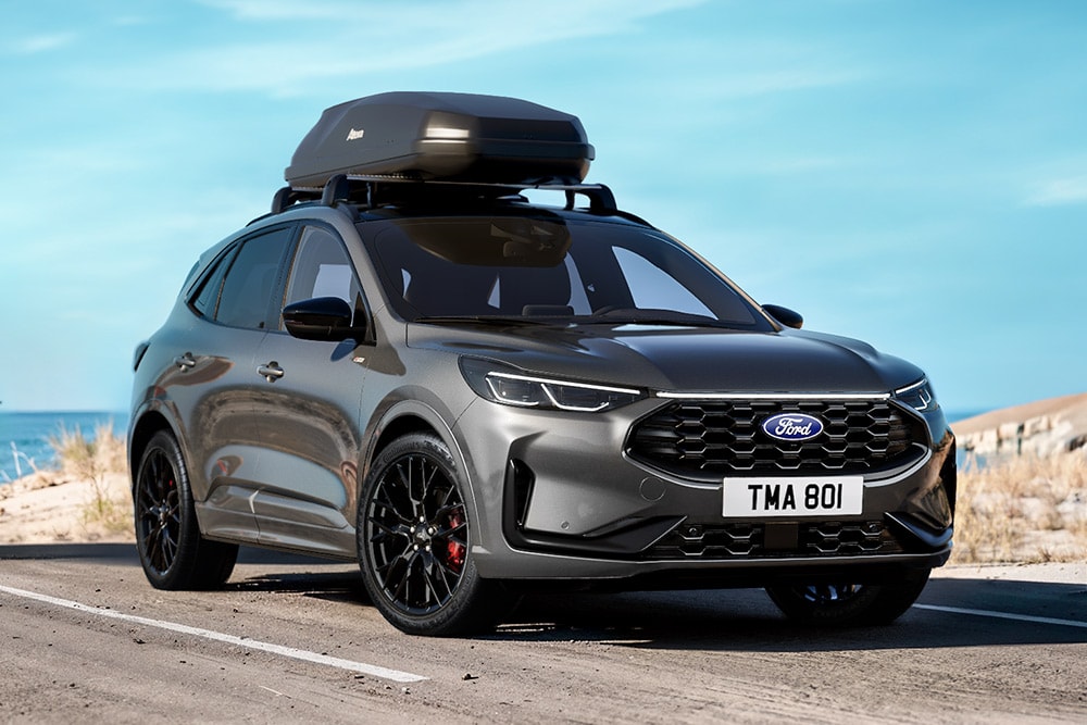 Pohled zepředu ze 3/4 na šedý Ford Kuga se střešním boxem, zaparkovaný na silnici s mořem v pozadí