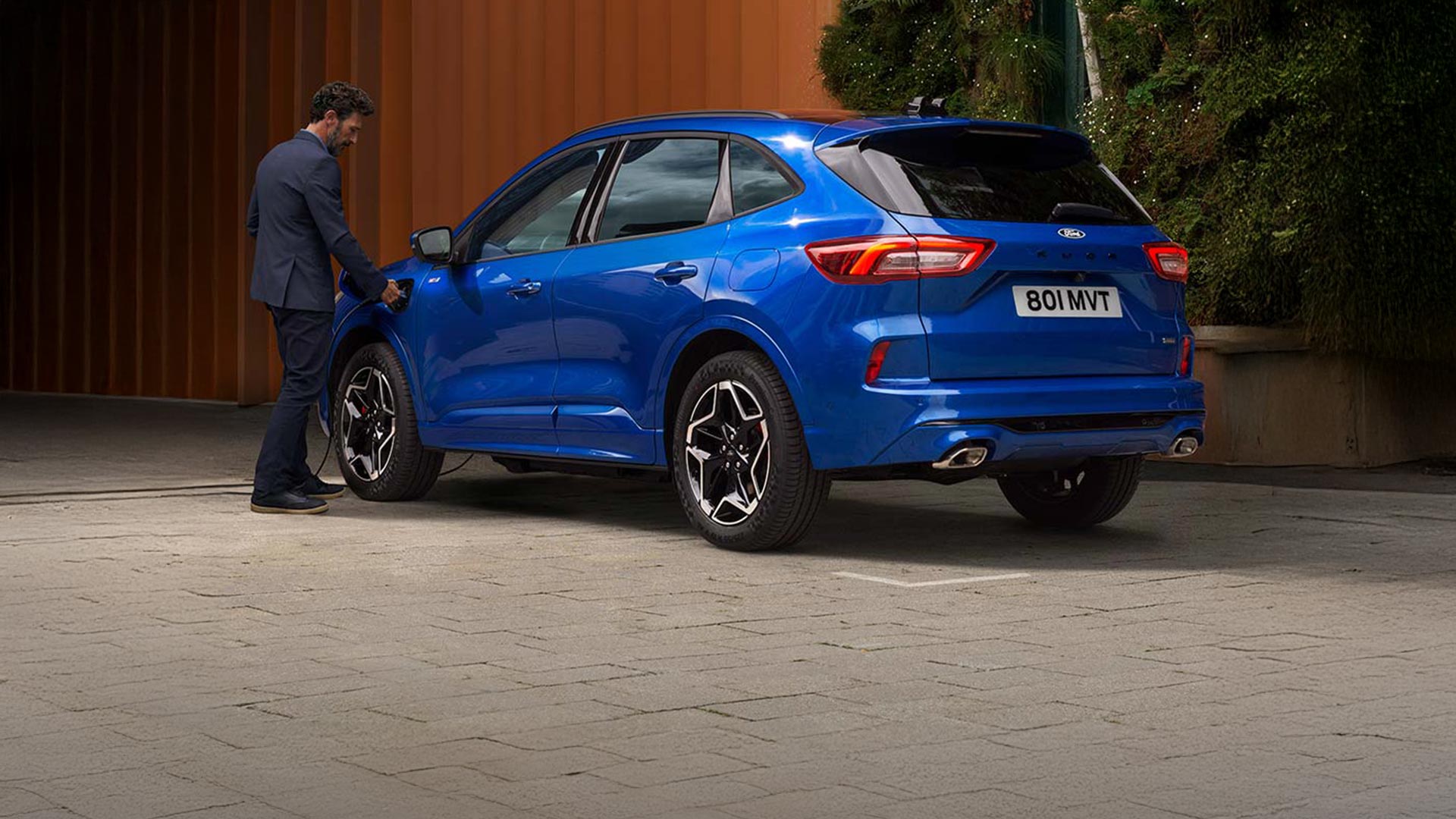 Rear view of a blue Ford Kuga being charged by a person in an urban setting.