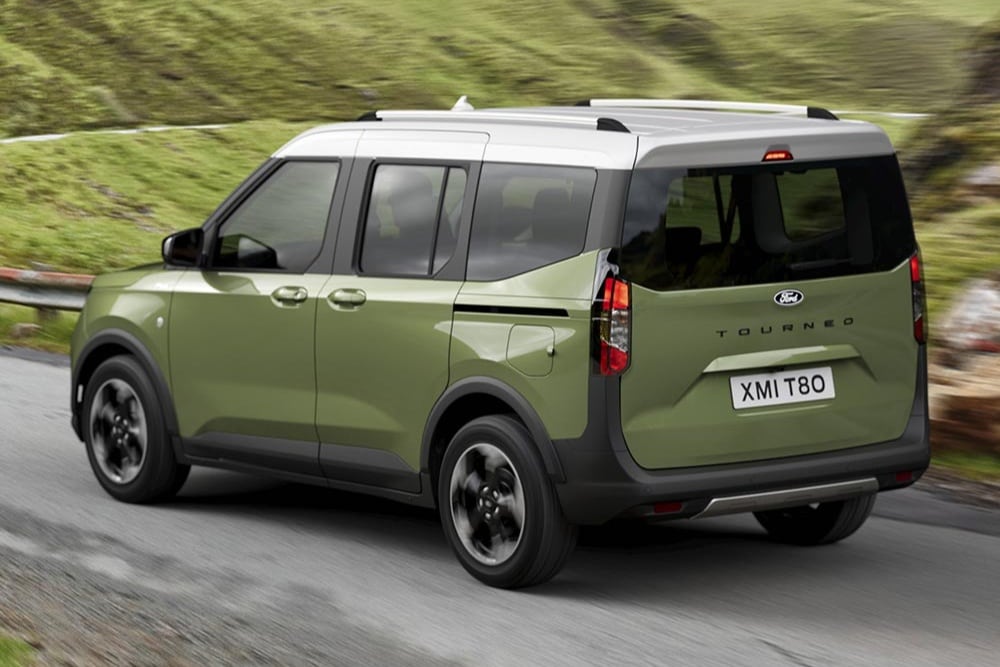 Zelený Ford Tourneo Courier je zachycen ze zadního šikmého pohledu, jak jede po klikaté silnici mezi zelenými kopci.