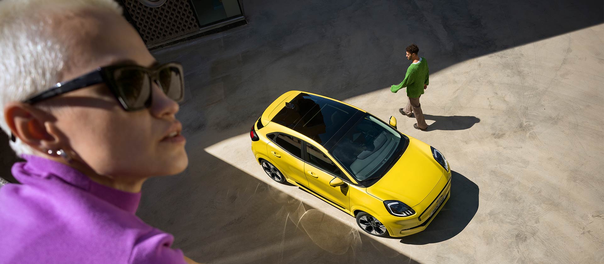 Pohled z ptačí perspektivy na žlutý elektrický Ford Puma Gen-E®. Člověk ve fialovém oblečení v popředí.