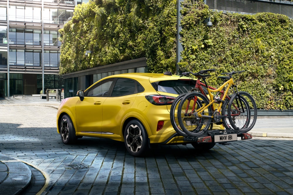 Žlutý Ford Puma Gen-E zaparkovaný v městském prostředí se dvěma jízdními koly na zadním nosiči.