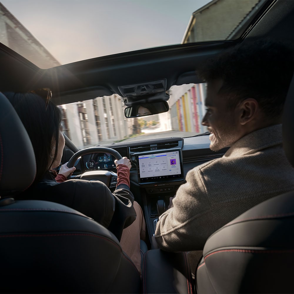 Řidič si povídá se spolujezdcem při jízdě sluncem zalitou krajinou s rezidenčními budovami.