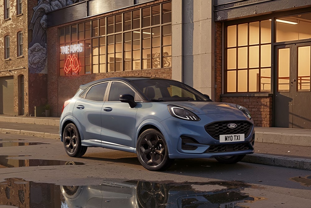 Modrý Ford Puma zaparkovaný na ulici ve městě při západu slunce. V kalužích se odrážejí budovy.