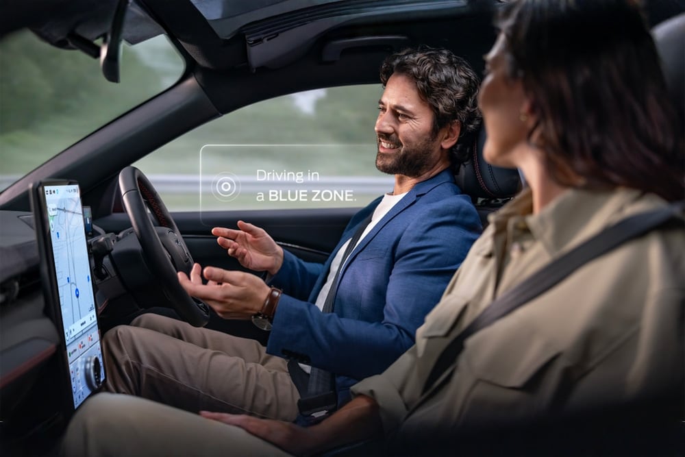 Muž a žena ve voze Mustang Mach-E využívají hands-free jízdy díky systému Ford BlueCruise. Upozornění říká "Jízda v BLUE ZONE“.