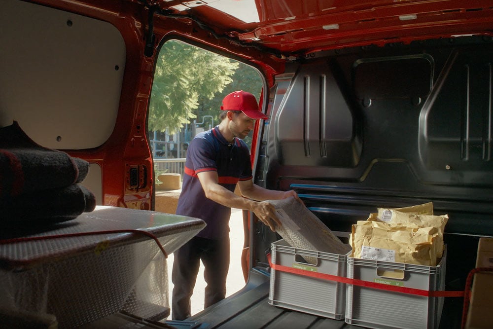 Kurýr vykládá balíky z červeného vozu Ford Transit Custom.