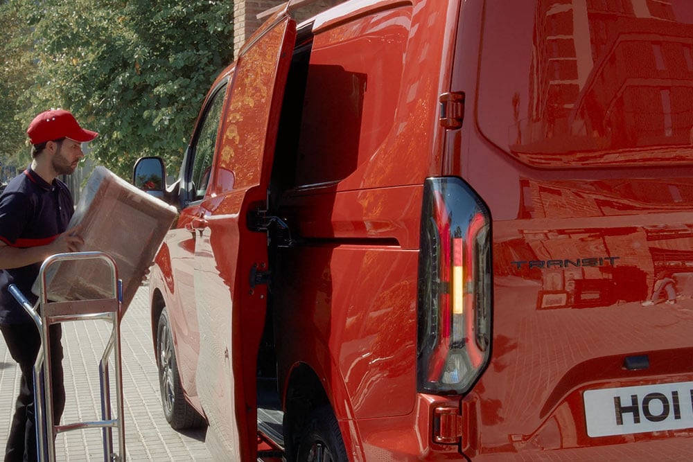 Kurýr vykládá balíky z červeného vozu Ford Transit Custom.