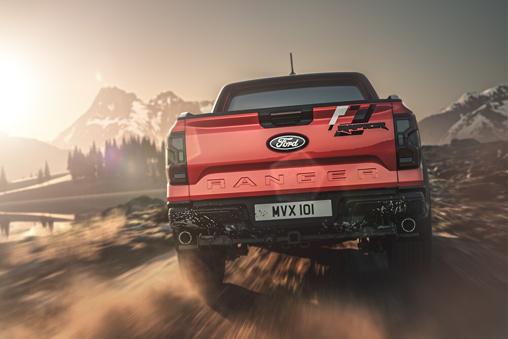 Pohled zezadu na červený Ford Ranger Raptor jedoucí po prašné cestě s horami v pozadí.