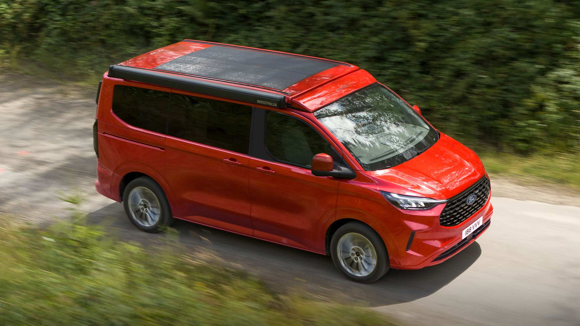 Červený obytný vůz Ford Transit Custom Nugget jedoucí po polní cestě.