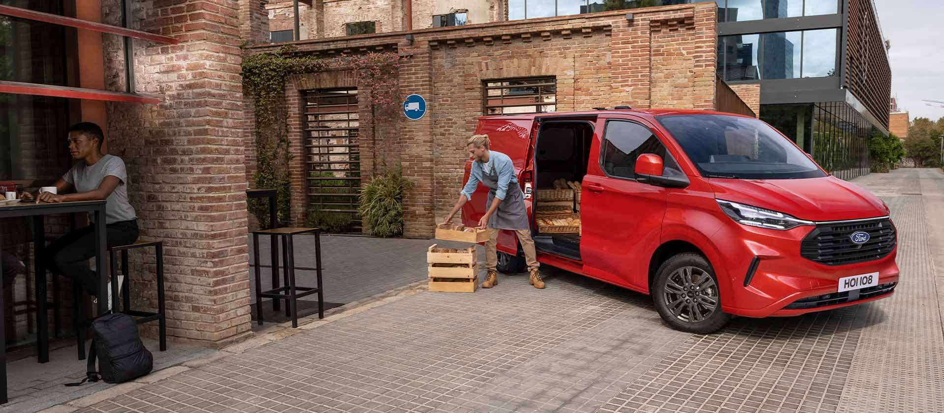 Člověk vykládá bedny z červené dodávky Ford Transit Custom zaparkované před kavárnou. 