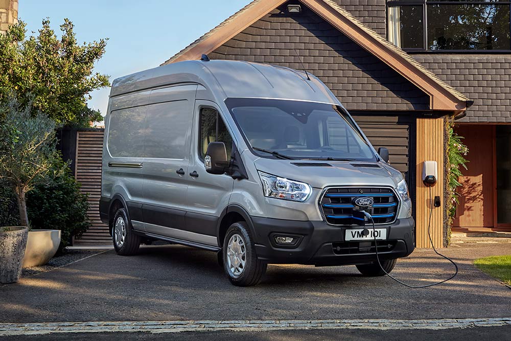 Stříbrná dodávka Ford E-Transit Van zaparkovaná na příjezdové cestě vedle domu a připojená k nabíjecí stanici.
