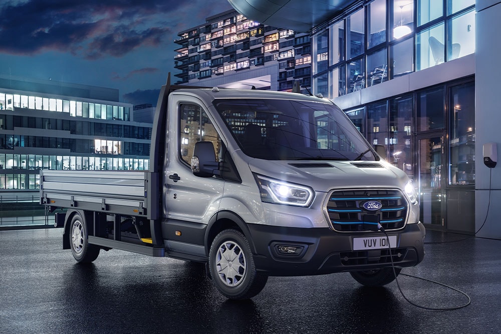 Stříbrný Ford E-Transit Podvozek s jednokabinou zaparkovaný v noci před moderní budovou.