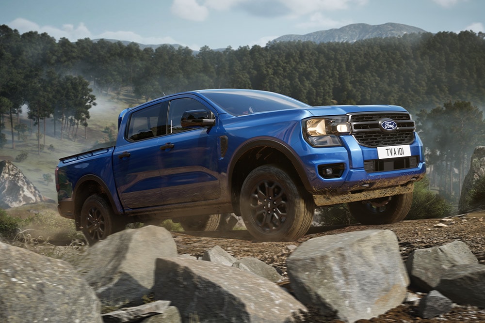Modrý pick-up Ford Ranger projíždí lesní cestou a zanechává za sebou prachovou stopu.