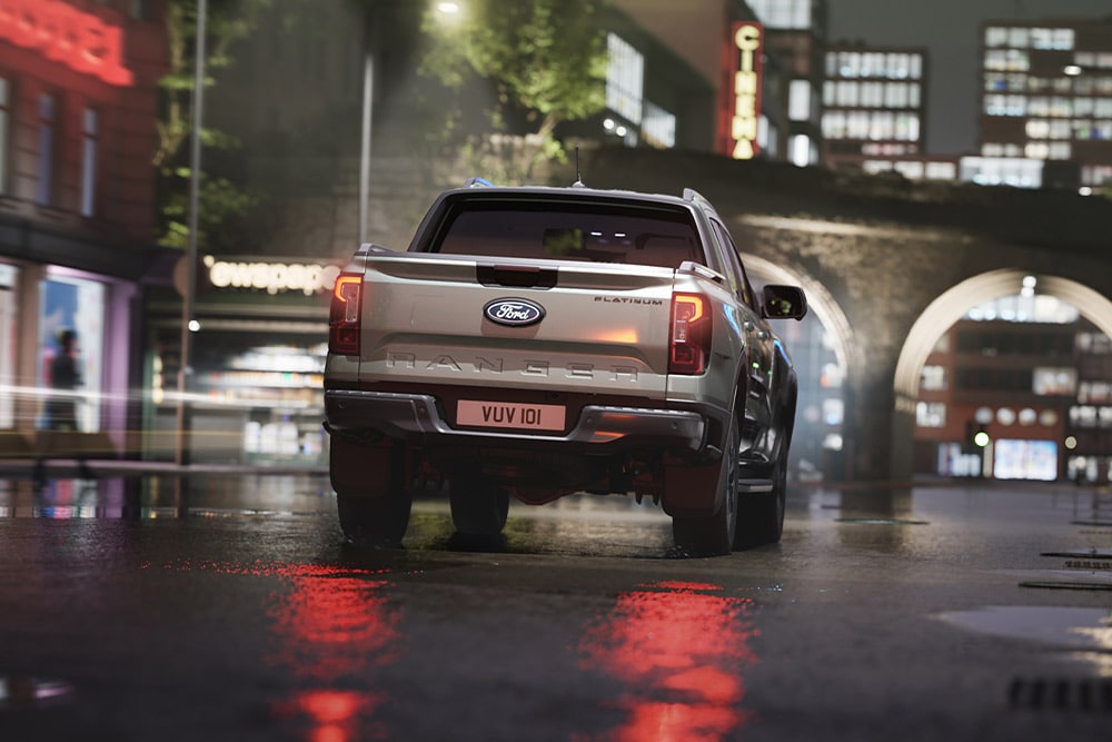 Zadní pohled na stříbrný Ford Ranger projíždějící v noci městskou scenérií ozářenou neony.