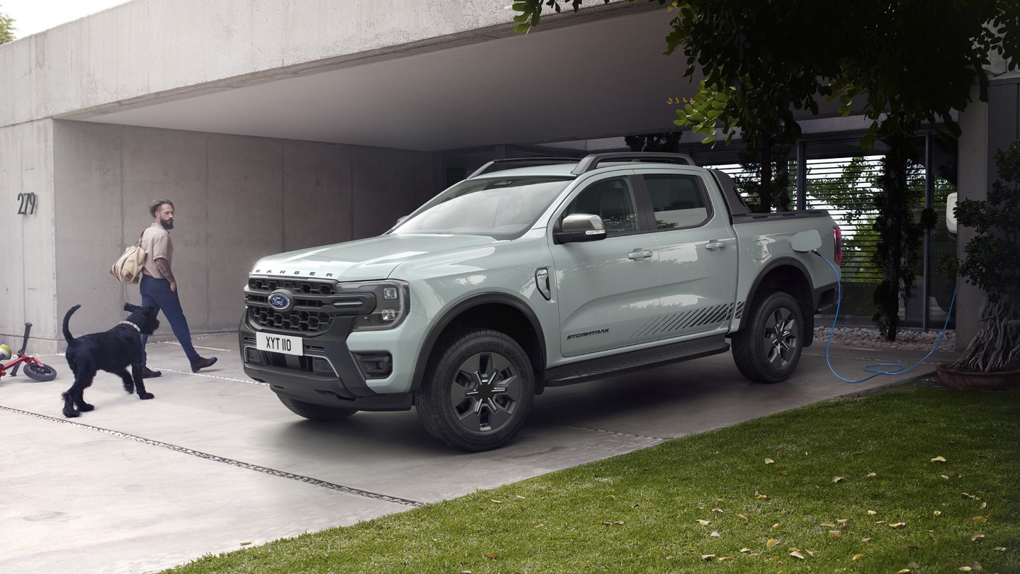 Muž se psem vchází do domu a míjí při tom plug-in hybridní Ford Ranger Stormtrak připojený do domácí nabíječky.