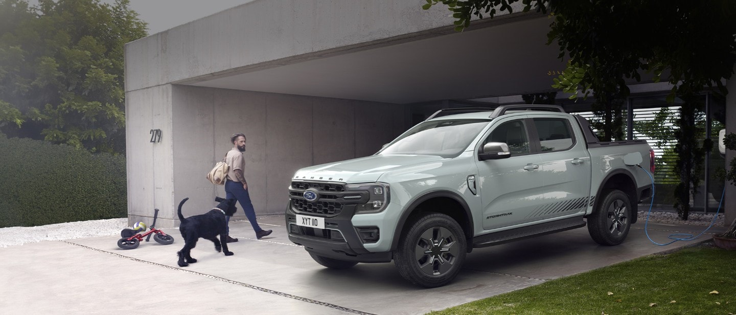 Muž se psem vchází do domu a míjí při tom plug-in hybridní Ford Ranger Stormtrak připojený do domácí nabíječky.