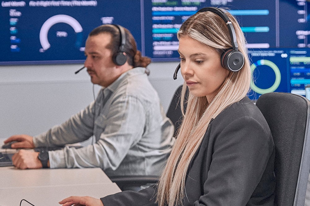  Dvě osoby se sluchátky pracují u počítačů v moderní řídicí místnosti.